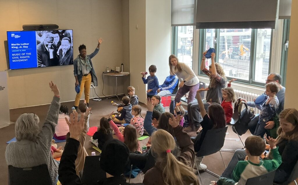 A view of a classroom from behind seated children and adults with an educator in front of a screen with her hand raised during a presentation. Participants also have their hands raised.