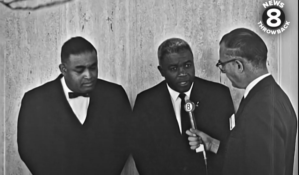 Screen grab of CBS 8 Interview with Jackie Robinson showing reporter holding a microphone in front of Jackie with another man standing to Robinson's right.